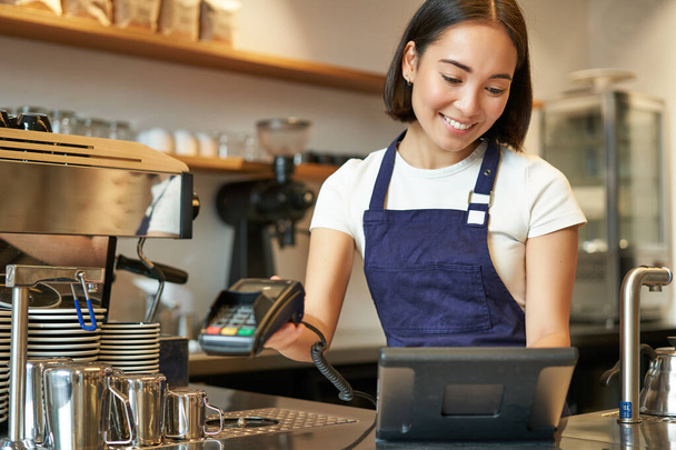 Gülümseyen Asyalı barista kız işlem sırası, POS terminaline girin, kahve dükkanında tezgahta çalışın.. - Fotoğraf, Görsel
