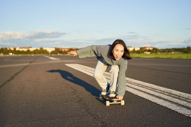 Beztroska dziewczyna na deskorolce, jeżdżąca na longboardzie po pustej drodze, trzymająca się za ręce bokiem i śmiejąca się. - Zdjęcie, obraz