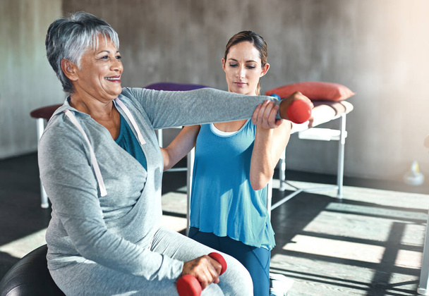 Предотвращение возрастной мышечной слабости с регулярной физиотерапией. пожилая женщина, использующая гири и фитнес-мяч с помощью физиотерапевта - Фото, изображение