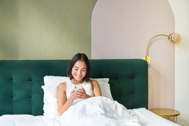 Yatakta uzanmış gülümseyen Asyalı kadının portresi, akıllı telefona bakıyor, mutlu yüzlü cep telefonu kullanıyor, tembel bir sabahta dinleniyor.. - Fotoğraf, Görsel