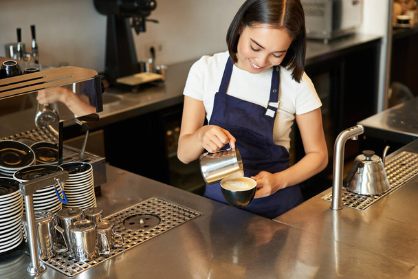 Улыбающаяся азиатка-бариста, персонал кафе наливает в кофе молоко, готовит капучино с искусством латте, стоит в синем фартуке за прилавком. - Фото, изображение