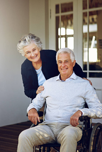 Σύντροφοι στη ζωή. Πορτρέτο ενός χαμογελαστού ηλικιωμένου σε αναπηρική καρέκλα και της συζύγου του έξω από το σπίτι τους - Φωτογραφία, εικόνα