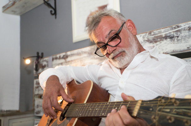 Vanhempi mies, jolla on valkoinen parta, istuu sängyllä soittamassa kitaraa. Miehellä on pyjama ja silmälasit. Vanhempien ja isovanhempien käsite. - Valokuva, kuva