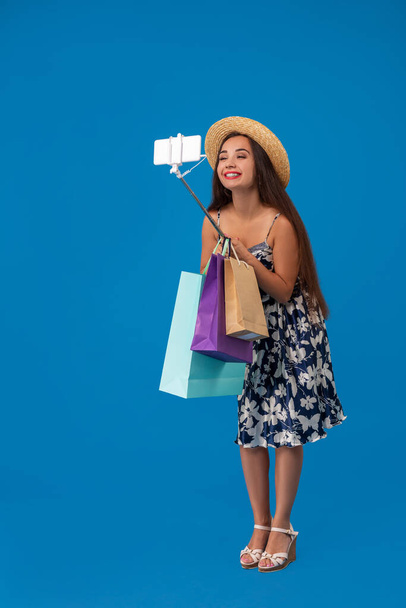 Trendy cheerful woman in casual clothes with shopping bags using selfie stick to take a self portrait on blue studio background with copy space. Emotions. Emotional woman - Photo, Image