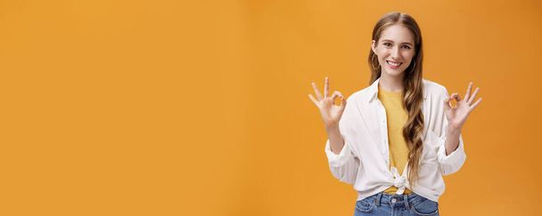 Estilo de vida. Indoor shot of assertive and assured charming stylish woman in blouse over t-shirt and accessories showing okay gesture with delighted self-assured smile giving being ok against orange wall - Foto, imagen