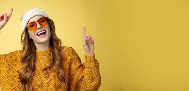 Girl enjoying party have fun smiling broadly happy gazing camera raising index fingers up dancing carefree laughing wearing sunglasses cozy sweater hat, celebrating weekends, yellow background. - Photo, Image