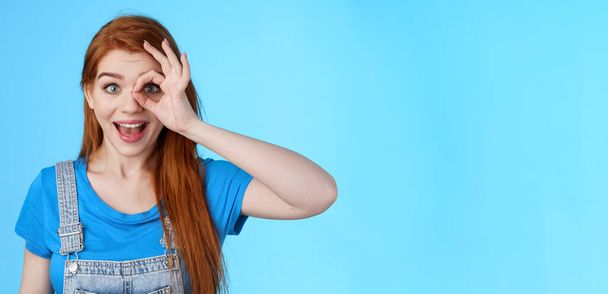 Dreamy curious attractive cheerful redhead woman surprised, look fascinated camera look through fingers okay sign, stand amused, gaze admiration blue background, check out promo. Copy space - Photo, Image