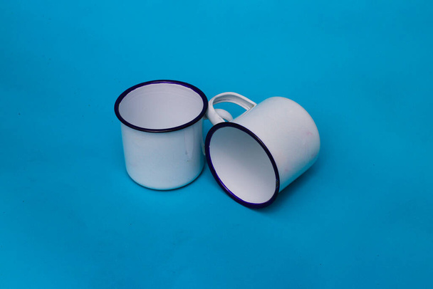 Dos tazas blancas sobre un fondo azul. Taza blanca esmaltada, taza de acero inoxidable, taza de camping. - Foto, imagen