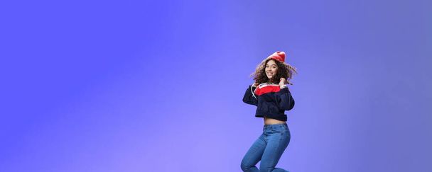Studio shot of cute girl with curly hair in beanie sweatshirt and sneakers jumping playful and carefree over blue background, having fun enjoying cool weather smiling broadly as flying in air. - Photo, Image