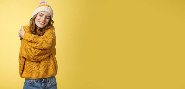 Tenderness, happiness wellbeing concept. Charming feminine cute stylish girl liking new warm sweater hugging embracing herself close eyes dreamy smiling feeling romantic coziness yellow background. - Photo, Image
