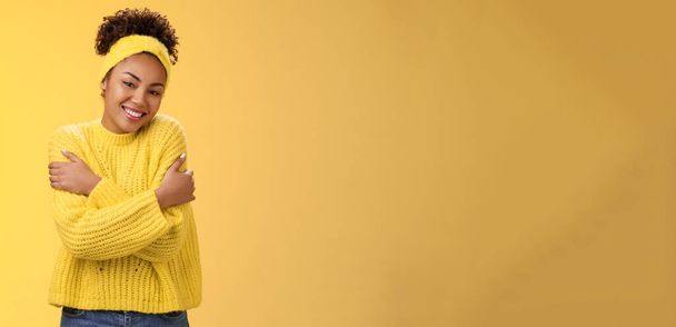 Encantadora mujer afroamericana femenina encantadora sonrisa tierna cabeza inclinada abrazándose a sí misma se siente comodidad amor suavemente abrazándose hombros usando cómodo suéter, sonriendo felizmente fondo amarillo. - Foto, imagen