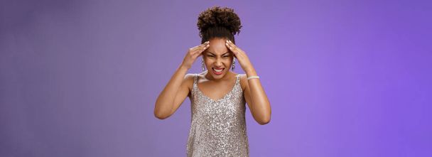 Pressured intense worried african-american woman in silver glittering dress grimacing cringing painful headache hold hand forehead suffer pain migraine, standing bothered blue background. - Photo, Image