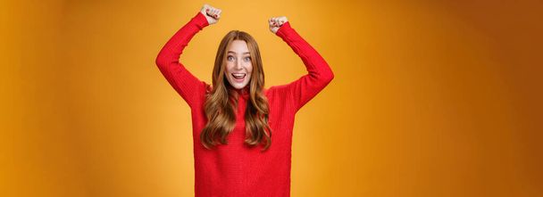 Optymistyczna szczęśliwa i wspierająca ruda dziewczyna krzyczy wiwatujące słowa, podnosząc ręce radośnie i uśmiechając się szeroko triumfując, świętując sukces i wygraną, pozując zadowolona i podekscytowana pomarańczową ścianą - Zdjęcie, obraz