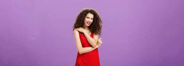 Schüchternes feminines, elegantes europäisches Weibchen mit lockiger Frisur und Lippenstift in rotem Luxuskleid, das die Schulter berührt, lächelt sanft über lustige Witze des Freundes beim romantischen Abendessen über lila Wand - Foto, Bild