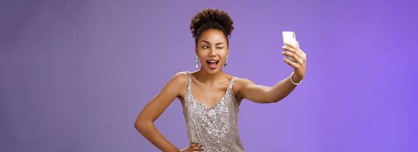 Élégant sassy afro-américaine b-day girl prenant selfie nouvelle élégante robe brillante argentée étendre bras tenant smartphone posant écran d'affichage clin d'oeil amusé avoir du plaisir à sourire largement, fond bleu. - Photo, image