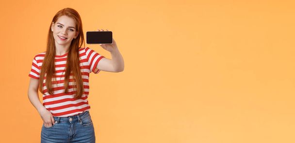 Уверенная в себе привлекательная девушка 20-х годов, девушка-имбирь, представляющая приложение для смартфона, рекомендующая классную игру, показывающая экран мобильного телефона хмурым, гордо улыбающимся, хвастающимся победным счетом, стоящим на оранжевом фоне. - Фото, изображение