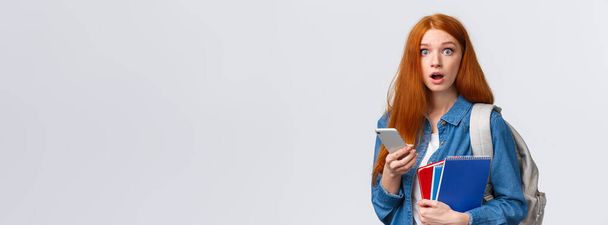 Freshman girl with red long hair, dont know where her class, looking questioned and confused, stare worried, holding smartphone, backpack and notebooks, clueless where go, white background. - Photo, Image