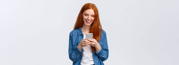 Chatten, communicatie en mobiel concept. Vrolijke roodharige glimlachende meisje sms 'en vriend, lachen als het kijken naar grappige vlog, op zoek smartphone display blij, staande witte achtergrond. - Foto, afbeelding