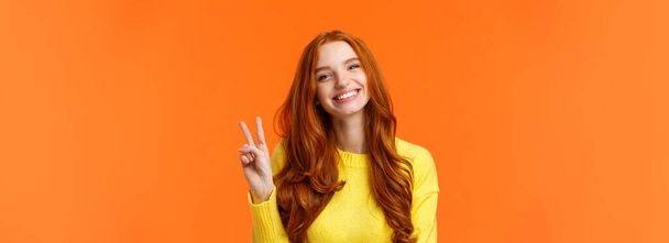 Uzun kıvırcık kızıl saçlı, belden aşağı neşeli bir kadın, barış işareti gösteriyor ve mutlu bir şekilde gülümsüyor, kış tatili gibi, turuncu arka planda duruyor.. - Fotoğraf, Görsel