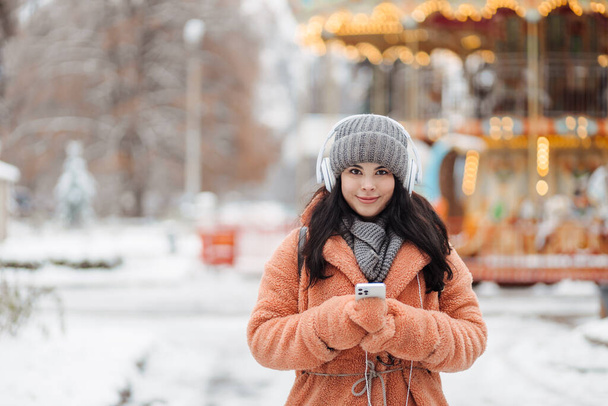 Csinos fiatal, hosszú hajú nő rózsaszín kabátban élvezze a téli napot és zenét hallgatni fejhallgatóval a havas parkban - Fotó, kép