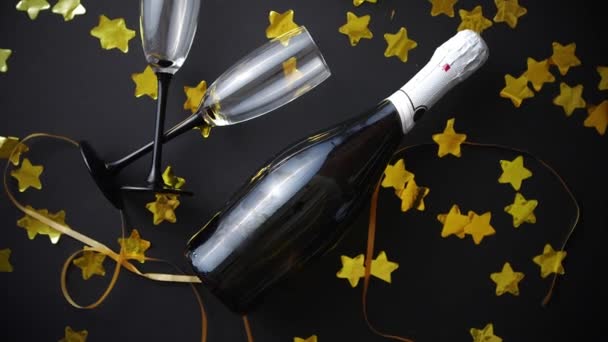 Siyah arka planda altın yılanlar ve konfeti bulunan bardaklar ve bir şişe şampanya. Yeni yıl veya parti kutlaması konsepti. Üst Manzara. Düz yatıyordu. - Video, Çekim