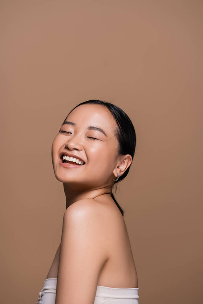 Joyful asian woman in top closing eyes while posing isolated on brown  - Zdjęcie, obraz