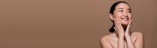 Positive asian woman with makeup and naked shoulders touching cheeks isolated on brown, banner  - Foto, Imagem