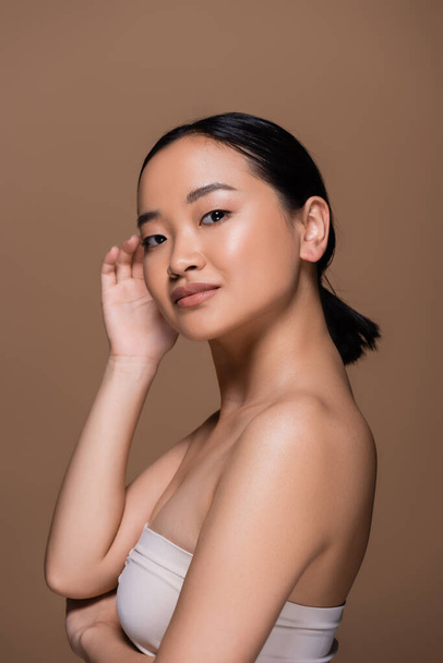 Young asian model with naked shoulder looking at camera isolated on brown  - Foto, Imagen