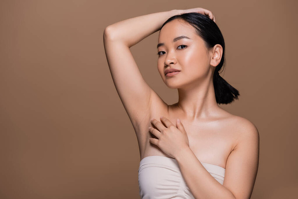 Pretty asian woman with naked shoulders touching fair and looking at camera isolated on brown  - Φωτογραφία, εικόνα