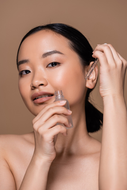 Asian woman with naked shoulders holding serum isolated on brown  - Photo, image