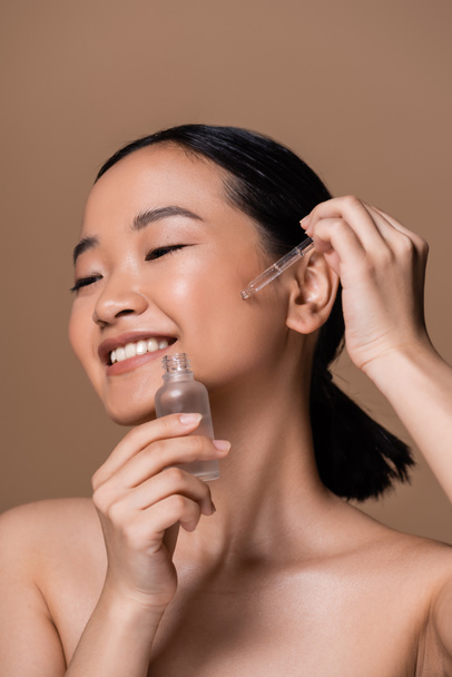 Positive asian model with naked shoulders applying serum on face isolated on brown  - Фото, зображення
