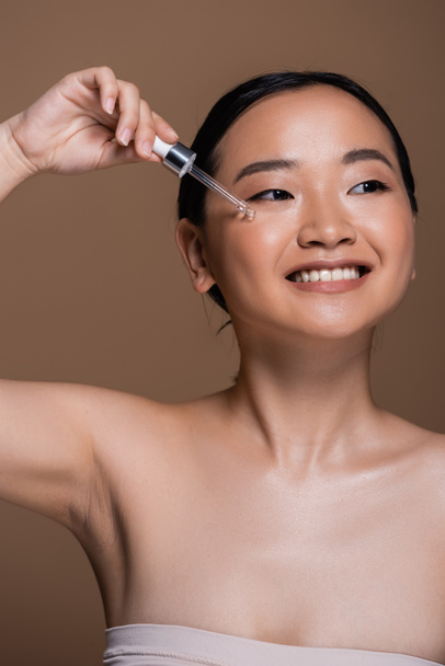 Smiling asian woman applying serum on face isolated on brown  - Фото, зображення