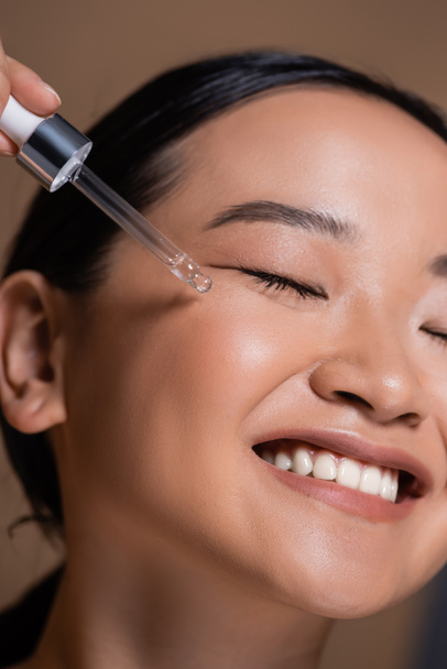 Close up view of smiling asian woman applying cosmetic serum on face isolated on brown  - Fotó, kép