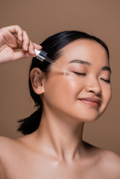 Joyful asian woman with dropper applying cosmetic serum on cheek isolated on brown  - Fotografie, Obrázek