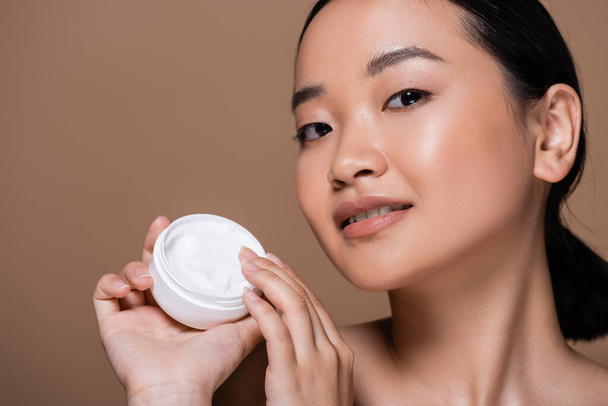 Young asian woman holding cosmetic cream and looking at camera isolated on brown  - Fotografie, Obrázek