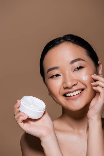 Positive asian woman with naked shoulders applying face cream isolated on brown  - Foto, Bild