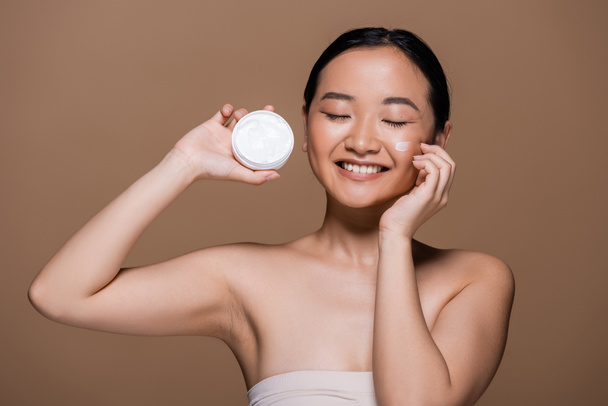 Positive asian woman with naked shoulders holding face cream isolated on brown  - Foto, Imagen