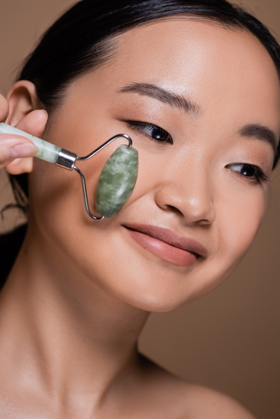 Portrait of asian woman holding jade roller near cheek isolated on brown  - Zdjęcie, obraz