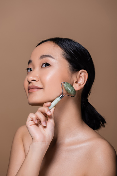 Young asian woman with naked shoulders holding jade roller and looking away isolated on brown  - Foto, imagen