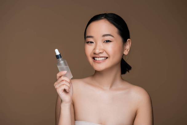 Positive asian woman with naked shoulders holding bottle of essential oil isolated on brown  - Foto, Imagen