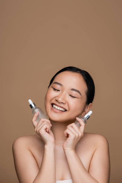 Pleased asian woman holding cosmetic serums isolated on brown  - Foto, Bild