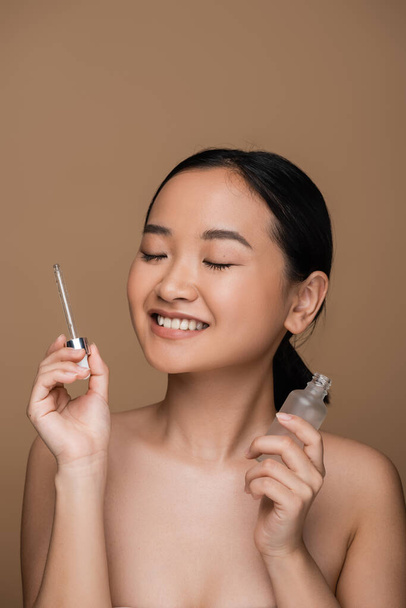Positive asian woman with naked shoulders holding serum isolated on brown  - Foto, afbeelding
