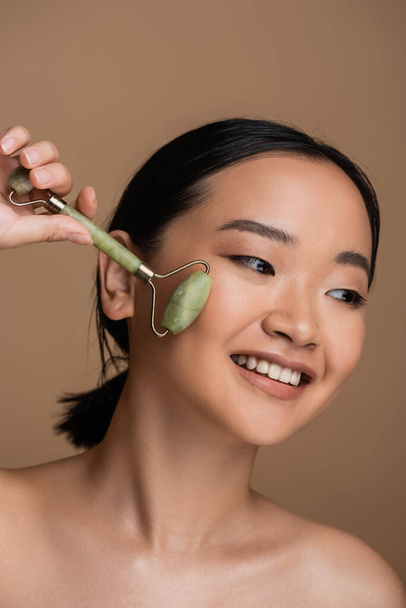 Positive asian model with naked shoulders using jade roller on face isolated on brown  - Fotografie, Obrázek