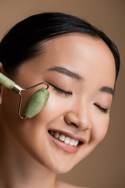 Pleased asian woman using jade roller on cheek isolated on brown  - Fotoğraf, Görsel