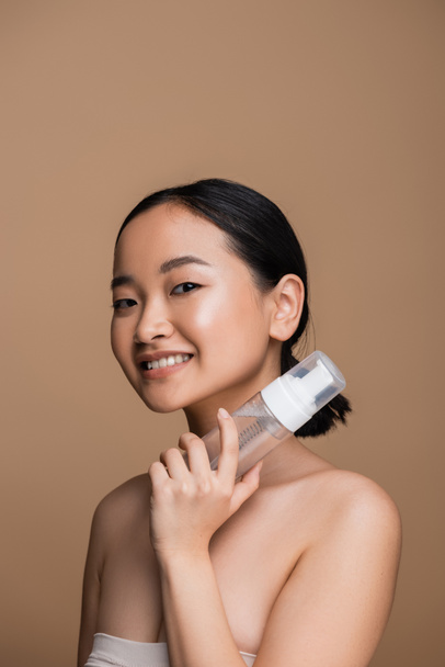 Молода азіатка з голими плечима тримає засіб для чищення обличчя ізольовано на коричневому
  - Фото, зображення