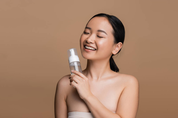 Positive asian model with naked shoulders holding cleansing foam isolated on brown  - Foto, Imagem