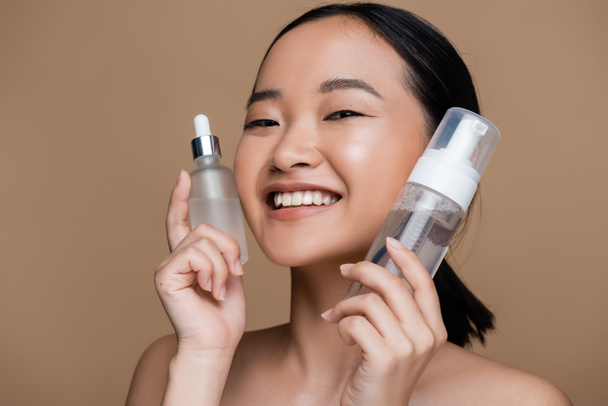 Positive asian woman with naked shoulders holding serum and face cleanser isolated on brown  - Photo, Image