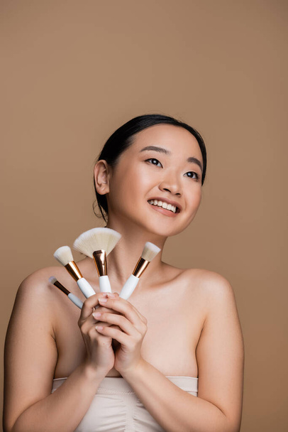 Positive asian model in top holding cosmetic brushes isolated on brown  - Photo, Image