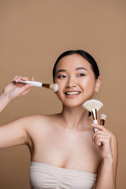 Young asian woman in top holding cosmetic brushes near face isolated on brown  - Photo, Image
