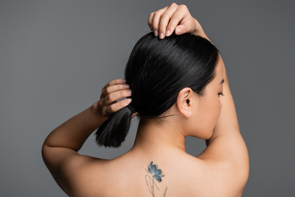 Side view of pretty asian woman with tattoo on back touching hair isolated on grey  - Foto, immagini